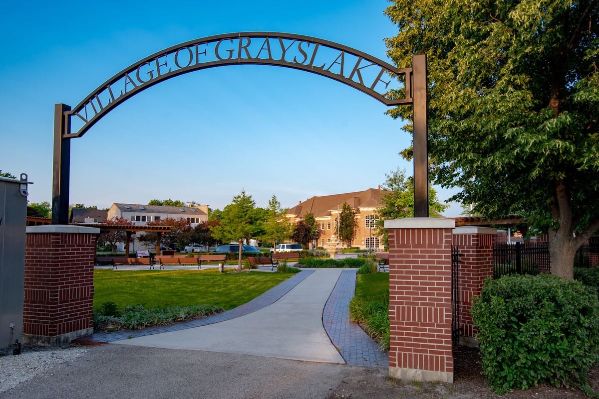 Wrought iron archway entrance with Village of Grays Lake sign at a park, leading to a paved pathway and open green space