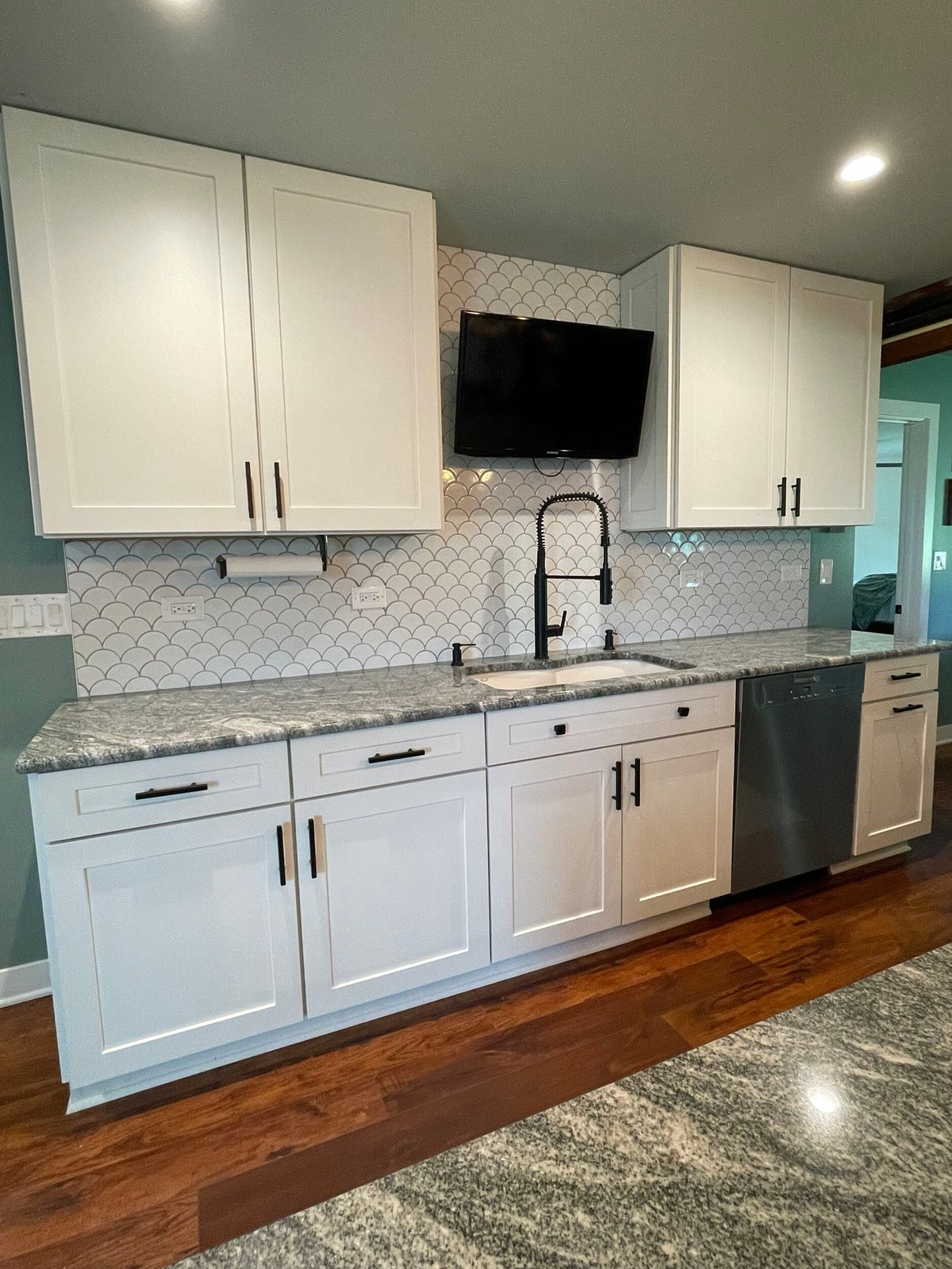 Modern kitchen with white cabinetry, granite countertops, geometric tile backsplash, and a flat-screen TV mounted above the sink area