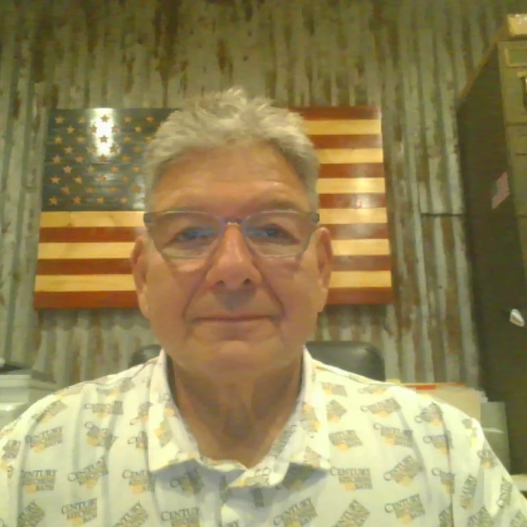 Older man with gray hair and glasses wearing a light patterned shirt, photographed indoors with an American flag visible in the background