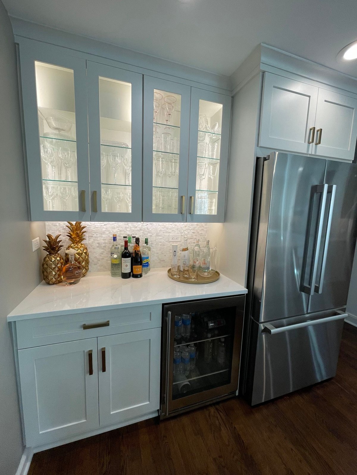 Modern kitchen with light blue cabinetry, white countertops, glass-front upper cabinets, stainless steel refrigerator, and wine bar with decorative items