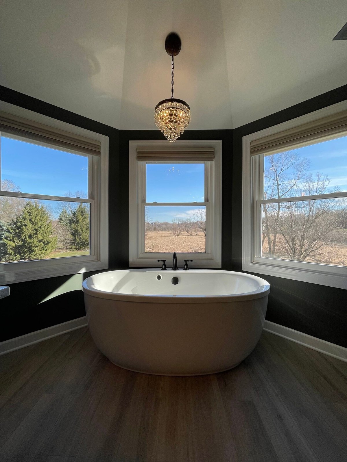 Modern freestanding white soaking tub in a spa bathroom with large windows overlooking rural landscape, black accent walls, and crystal chandelier