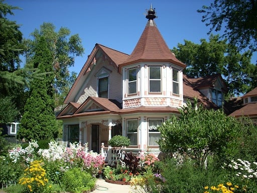 Victorian-style house with distinctive red turret, white trim, and colorful garden with blooming flowers under blue sky