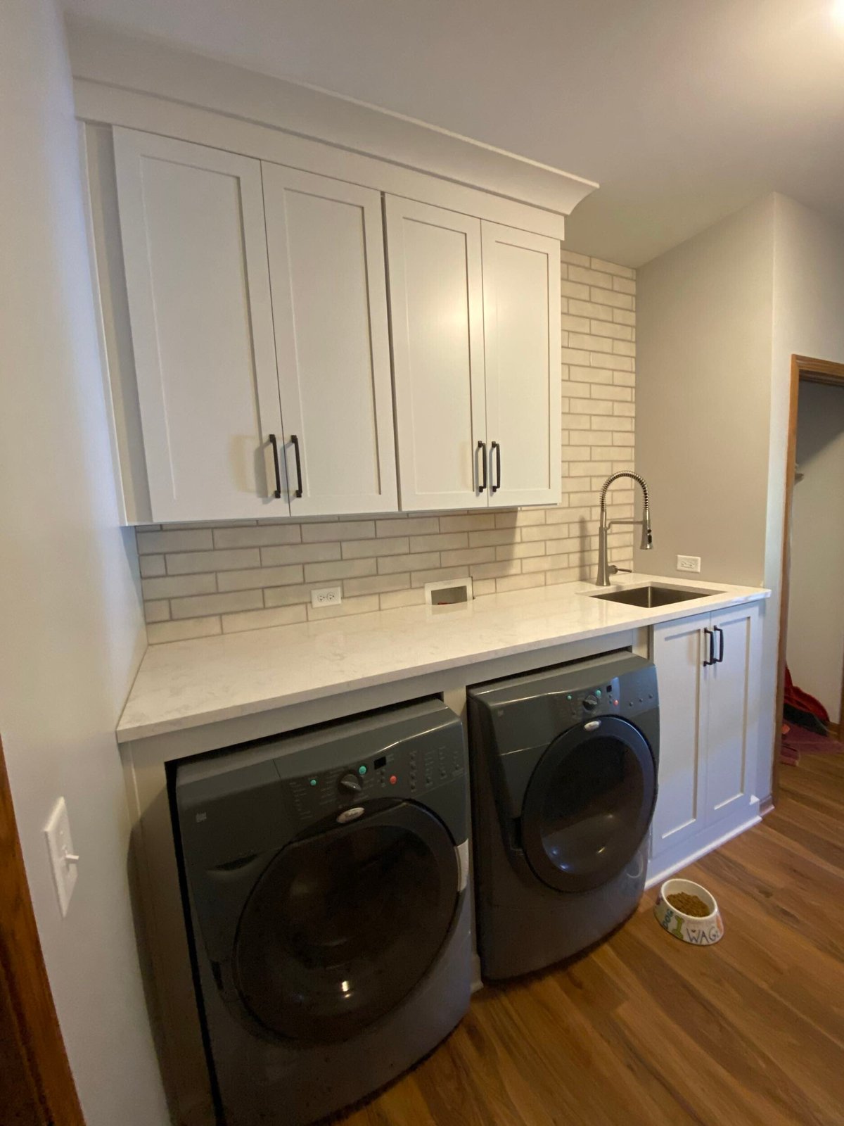 Modern laundry room with white cabinetry, white subway tile backsplash, quartz countertop, sink, and two black front-load washing machines on wooden floor
