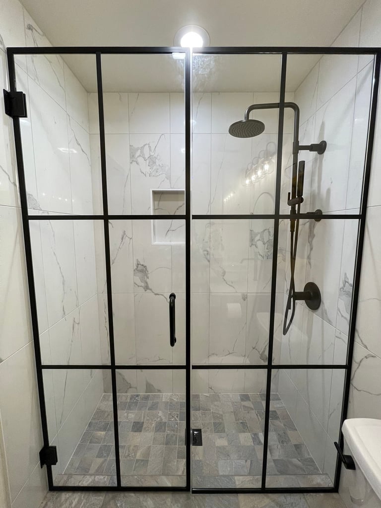 Modern glass shower enclosure with black metal frame, marble-look tile walls, rainfall showerhead, and patterned floor tile