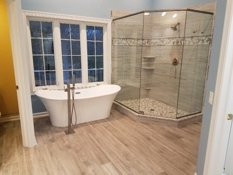 Modern luxury bathroom with freestanding white bathtub, glass-enclosed shower with tiled walls, wooden flooring, and large window with grid panes