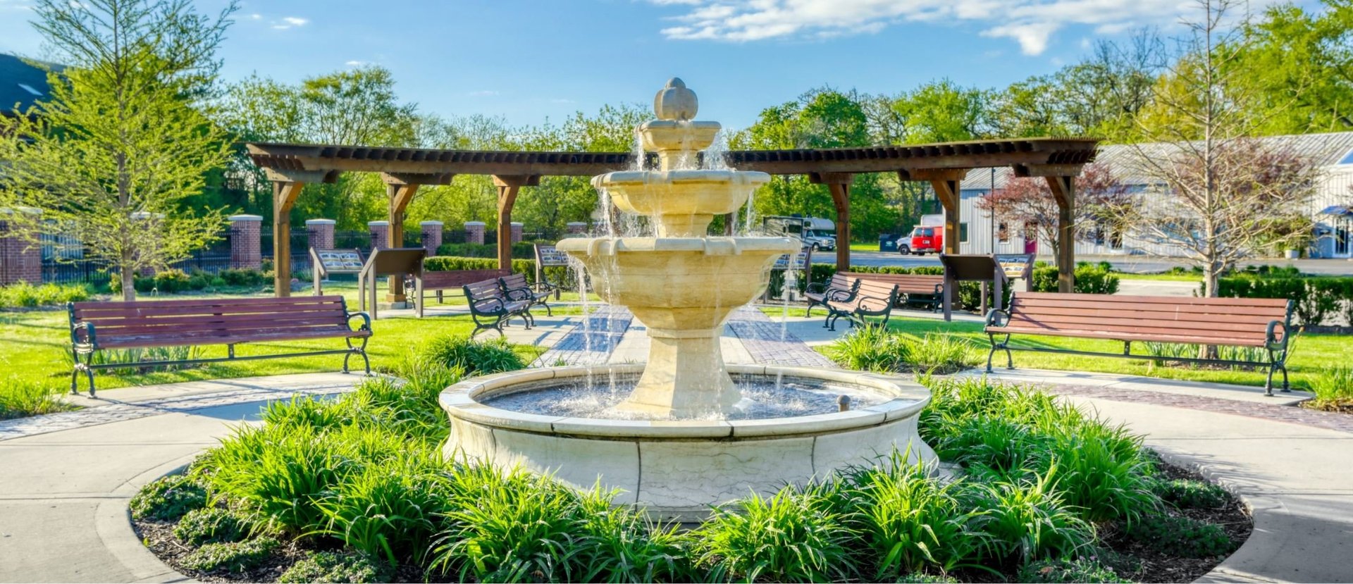 A decorative tiered fountain in a public park with benches, green landscaping, and a yellow building in the background