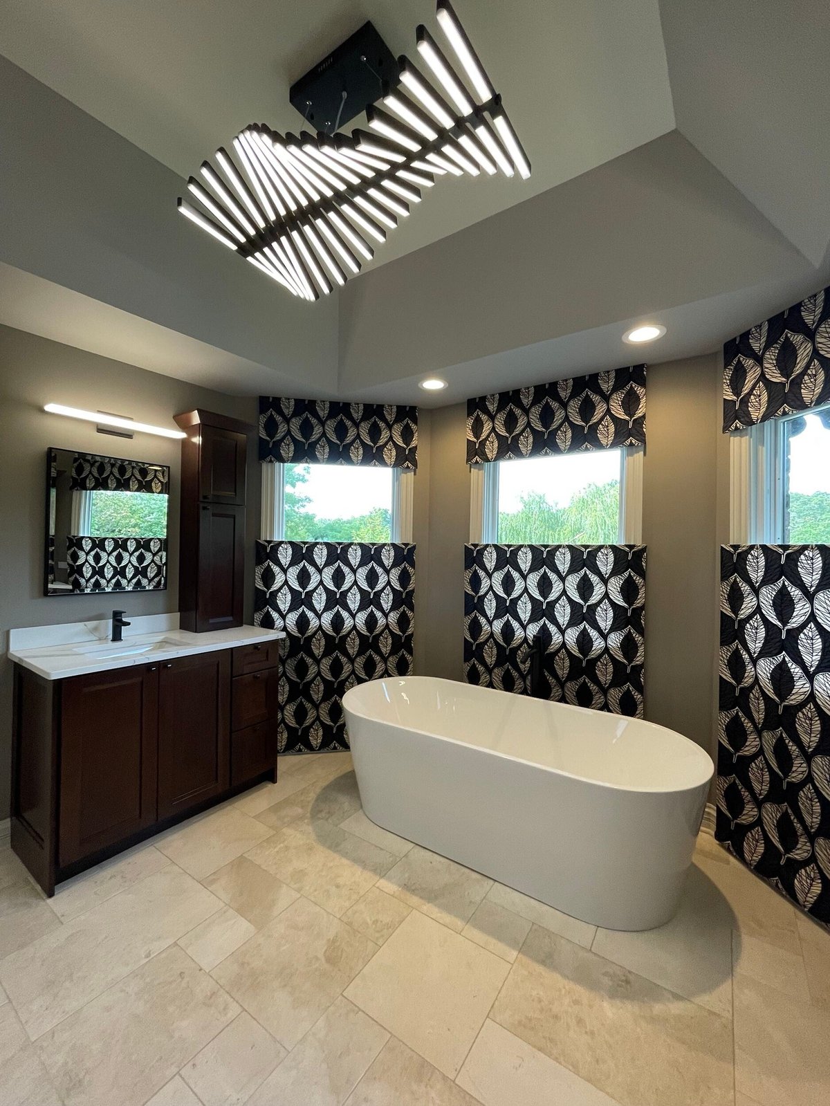 Modern luxury bathroom with white oval freestanding tub, dark wood vanity, geometric patterned curtains, and contemporary ceiling lighting