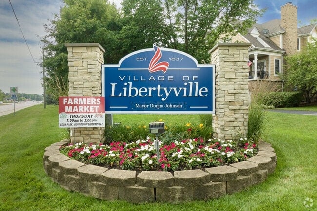 Village of Libertyville welcome sign with stone pillars and colorful flower bed in front of residential house