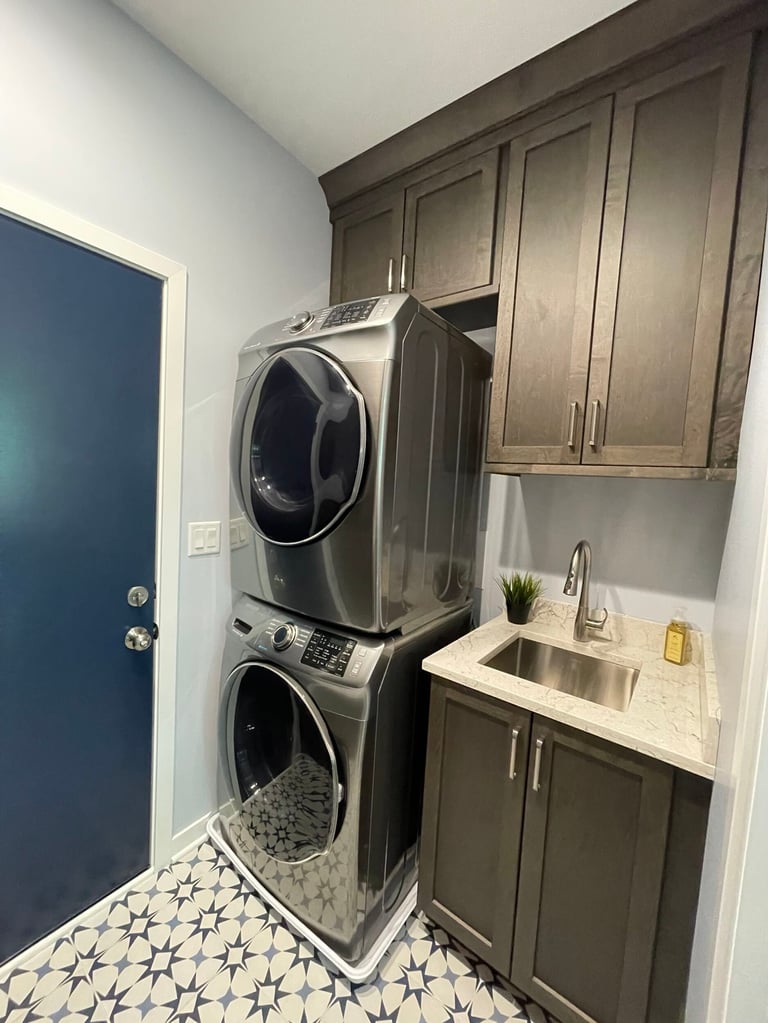 Modern laundry room with stacked washer and dryer, dark cabinetry, sink, and blue door